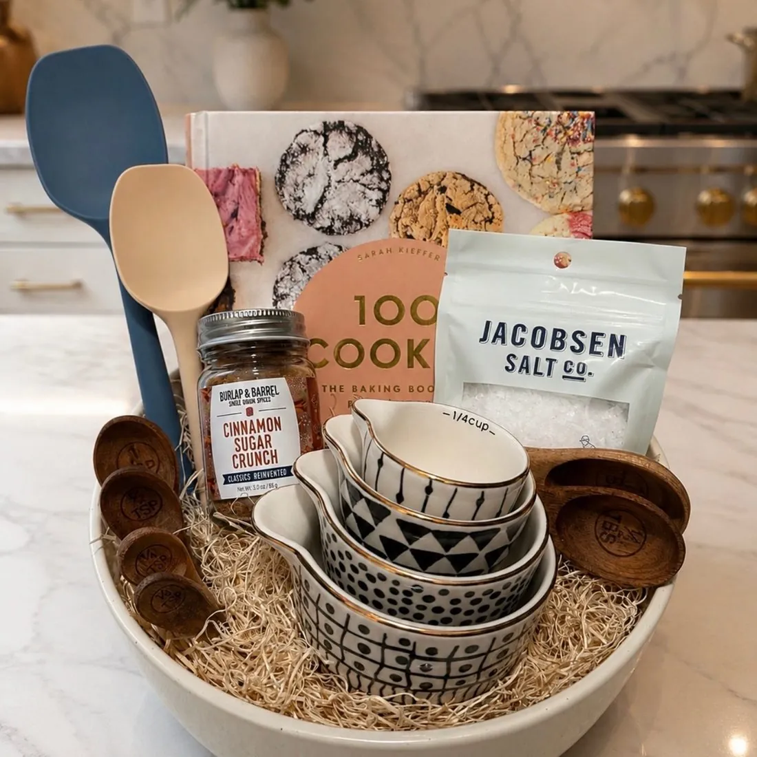 Baker's gift set arranged in a large ceramic bowl on a marble counter with the 100 Cookies cookbook, Burlap & Barrel Cinnamon Sugar Crunch, Jacobsen Salt Co. flake salt, black-and-white gold-rimmed measuring cups, wooden measuring spoons, and silicone spatulas