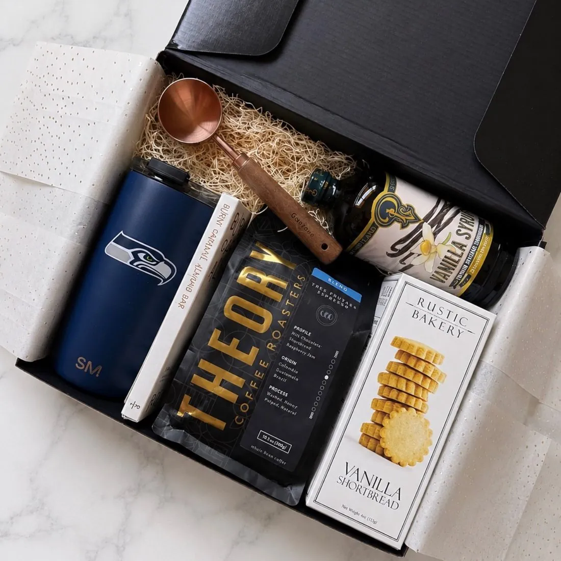 Open black gift box on white marble with a navy Seattle Seahawks tumbler, Theory Coffee Roasters coffee, vanilla syrup, Rustic Bakery vanilla shortbread, a copper coffee scoop, and a burnt caramel almond bar