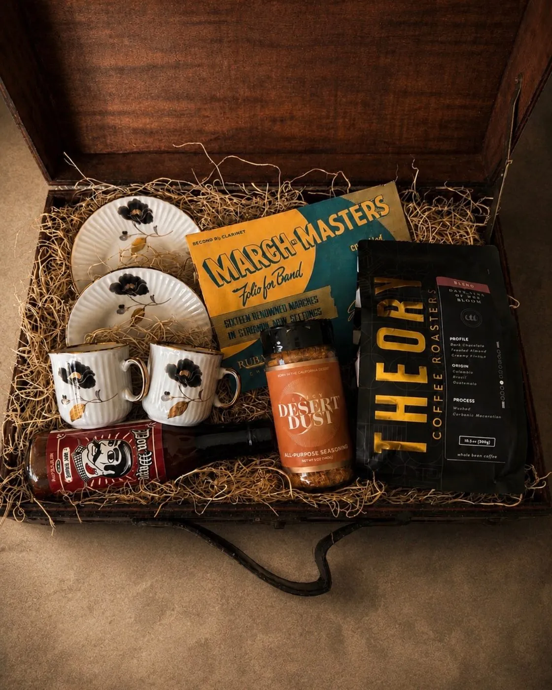 Vintage trunk gift box on straw filler with two black-floral gold-rimmed mugs and saucers, a vintage March-Masters band folio, Theory Coffee Roasters coffee, Desert Dust all-purpose seasoning, and a bottle of hot sauce