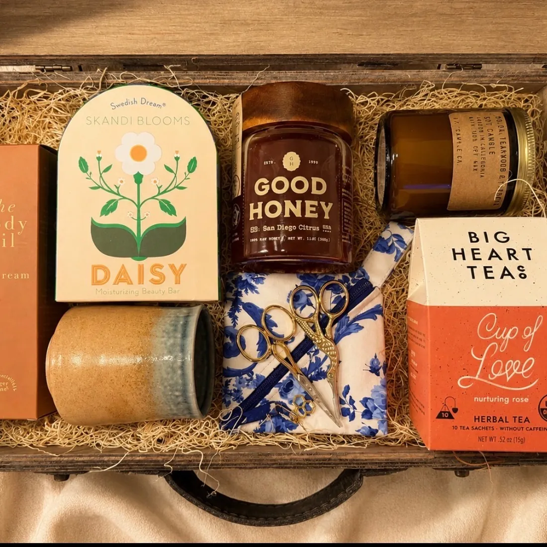 Wooden keepsake trunk filled with a Skandi Blooms Daisy beauty bar, a jar of Good Honey, an amber soy candle, The Body Oil in daydream, a stoneware tumbler, gold stork embroidery scissors on blue floral fabric, and a Big Heart Tea Cup of Love herbal tea box