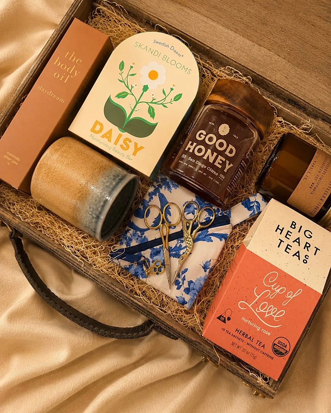 Open wooden keepsake trunk on cream fabric holding a Skandi Blooms Daisy beauty bar, a jar of Good Honey, an amber soy candle, The Body Oil in daydream, a stoneware tumbler, gold stork embroidery scissors on blue floral cloth, and a Big Heart Teas Cup of Love herbal tea box