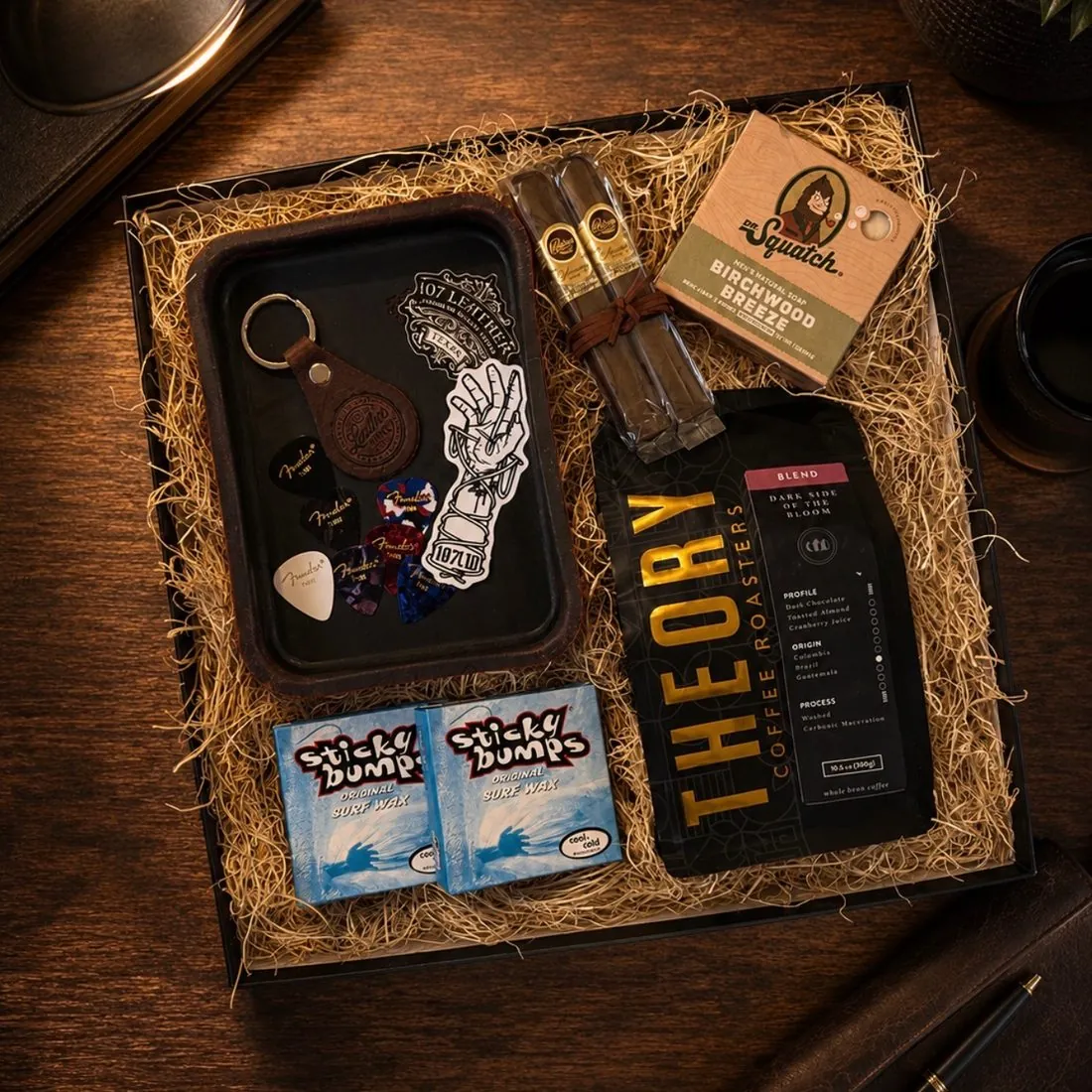 Overhead men's gift box on a dark wood desk with Fender guitar picks and a leather keychain on a 107 Leather tray, two cigars, Dr. Squatch Birchwood Breeze soap, two Sticky Bumps surf wax bars, and a bag of Theory Coffee Roasters whole bean coffee