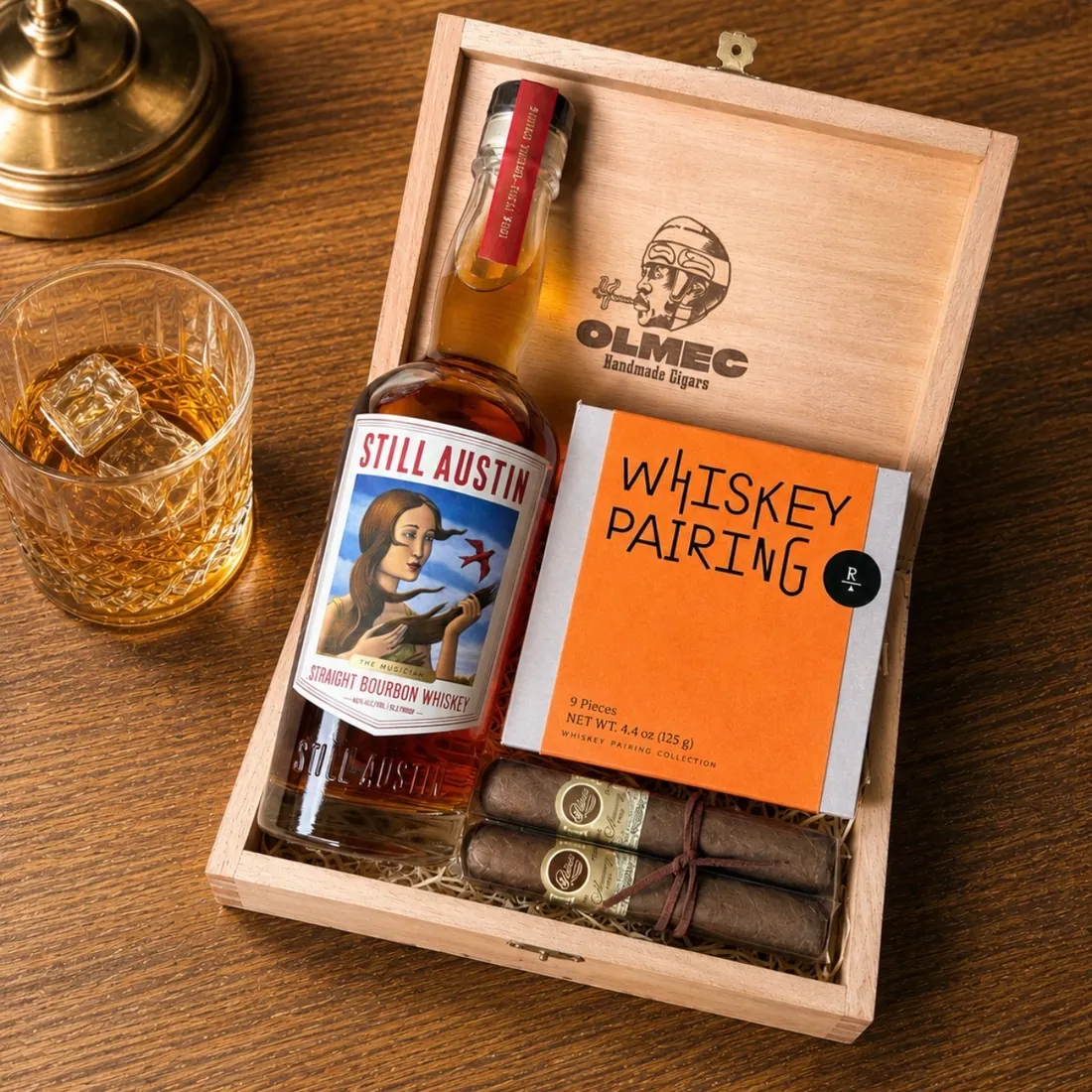 Whiskey-lover's gift set in an Olmec Handmade Cigars wooden box with a bottle of Still Austin Straight Bourbon Whiskey, a Whiskey Pairing sweets box, two cigars tied with twine, and a crystal glass of whiskey over ice alongside