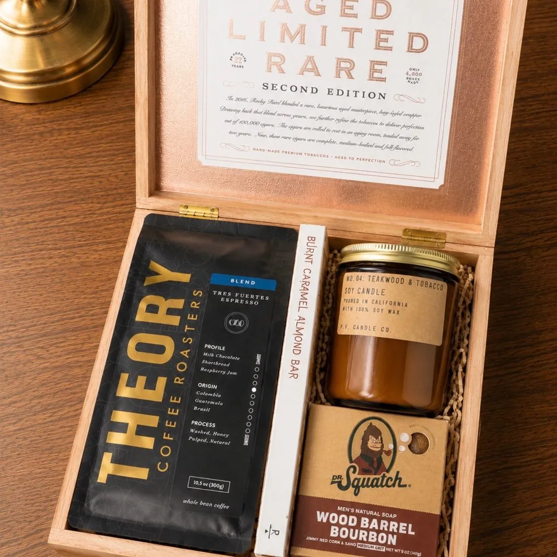 Open wooden Rocky Patel cigar box on a wood table holding Theory Coffee Roasters coffee, a burnt caramel almond bar, a P.F. Candle Co. teakwood and tobacco soy candle, and Dr. Squatch Wood Barrel Bourbon soap