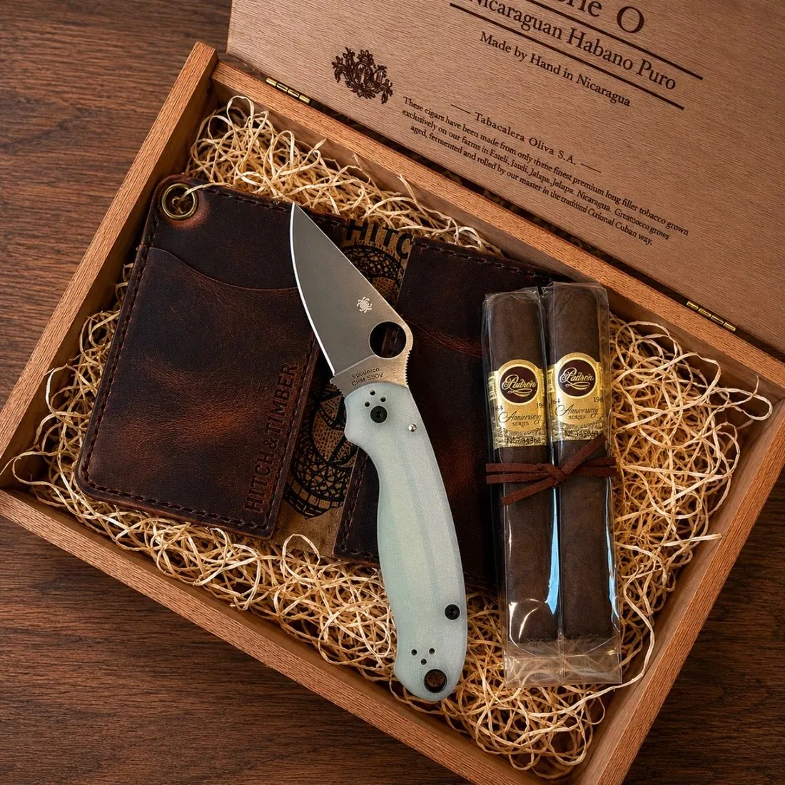 Open wooden cigar box on wood shavings with a Hitch & Timber leather card wallet, a Spyderco folding knife with a jade handle, and two Padrón Anniversary Series cigars