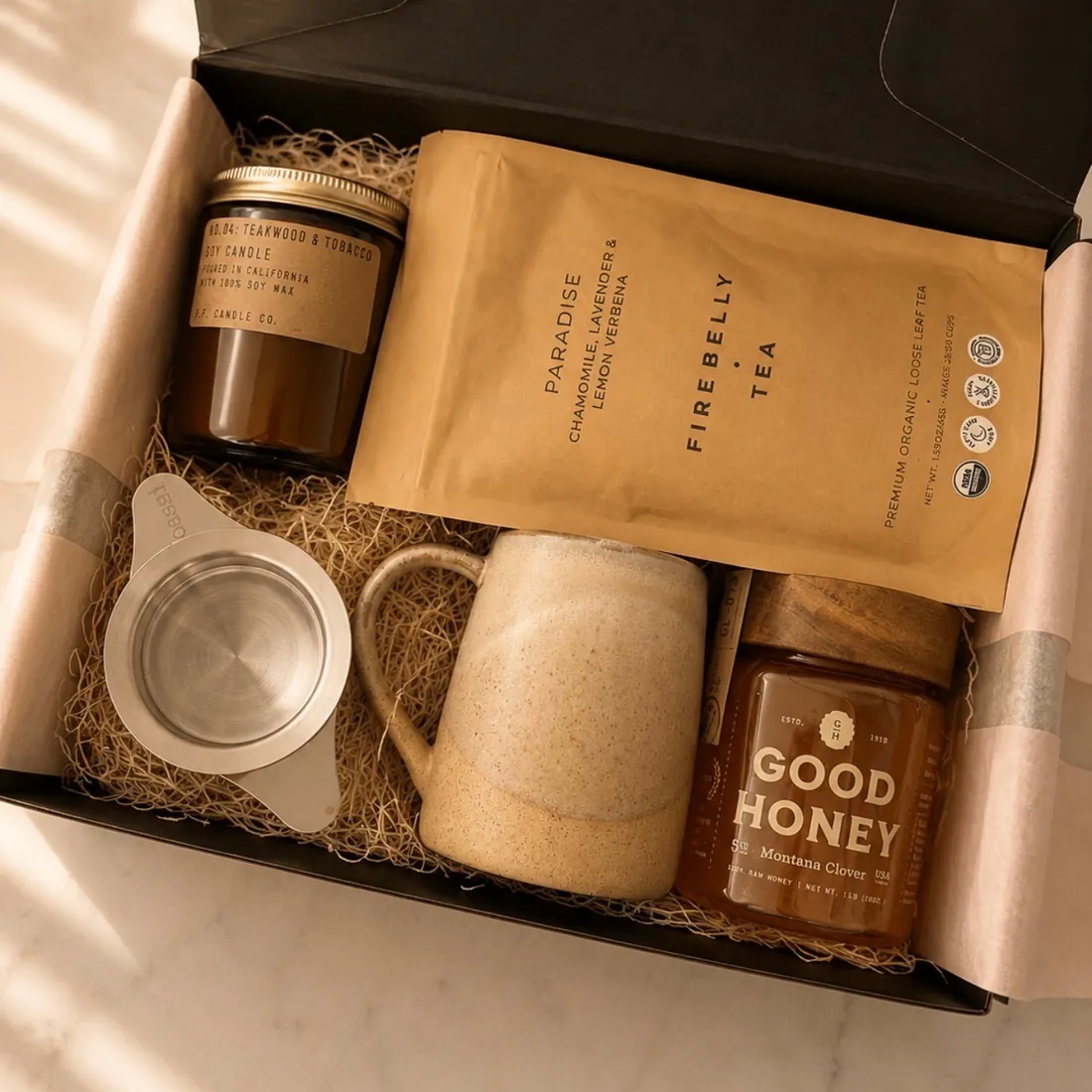 Black gift box lined with blush tissue paper holding a P.F. Candle Co. teakwood and tobacco soy candle, a bag of Firebelly Tea Paradise loose leaf tea, a cream stoneware mug, a stainless steel tea strainer, and a jar of Good Honey Montana Clover